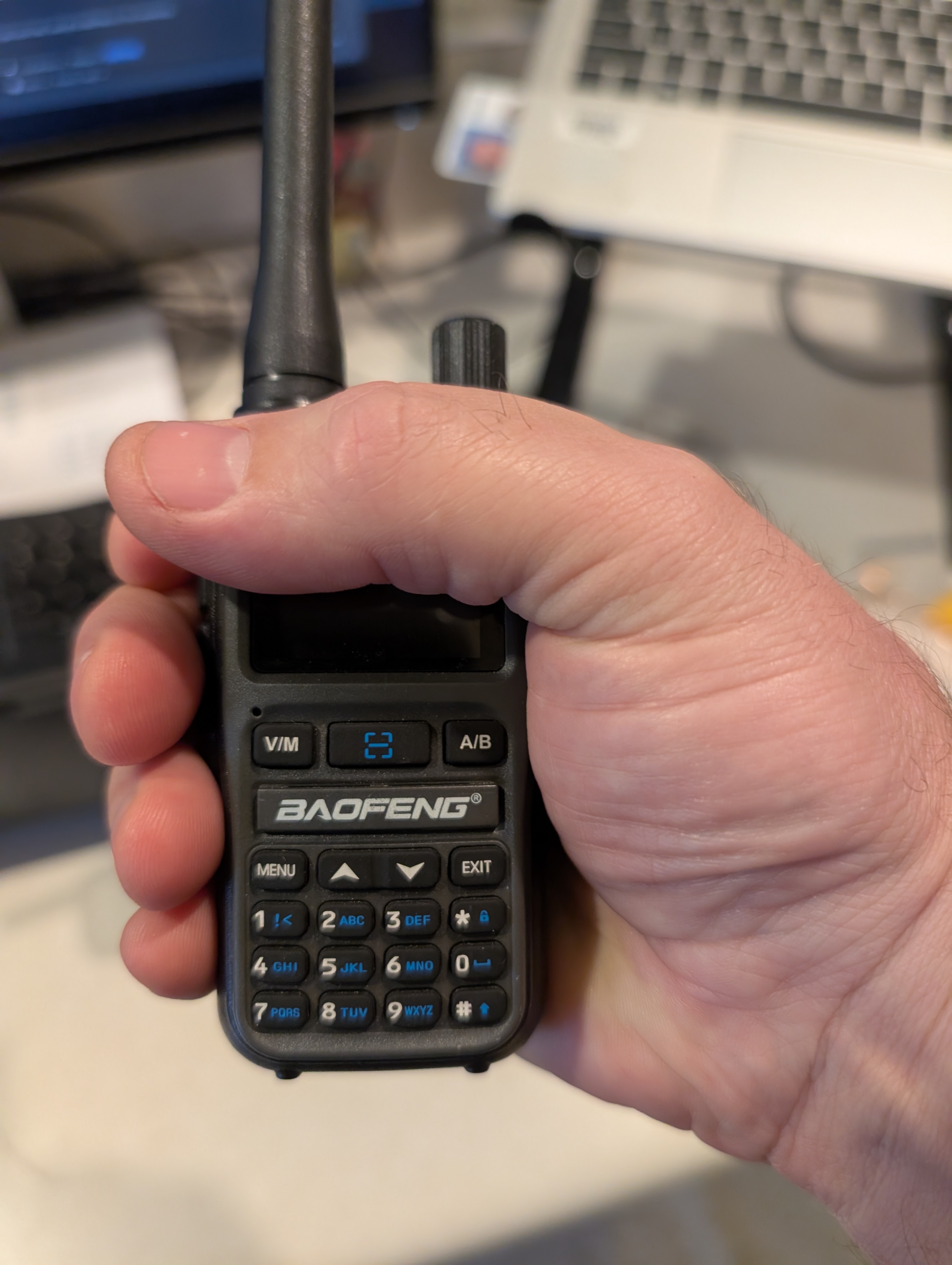 A picture of a Baofeng UV-5R mini, showing the authors thumb reaching past the edge of the radio. It's very compact, arguably too compact for many users hands.